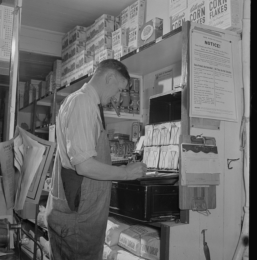 A Vermont general store owner penciling figures at his cash box in 1942