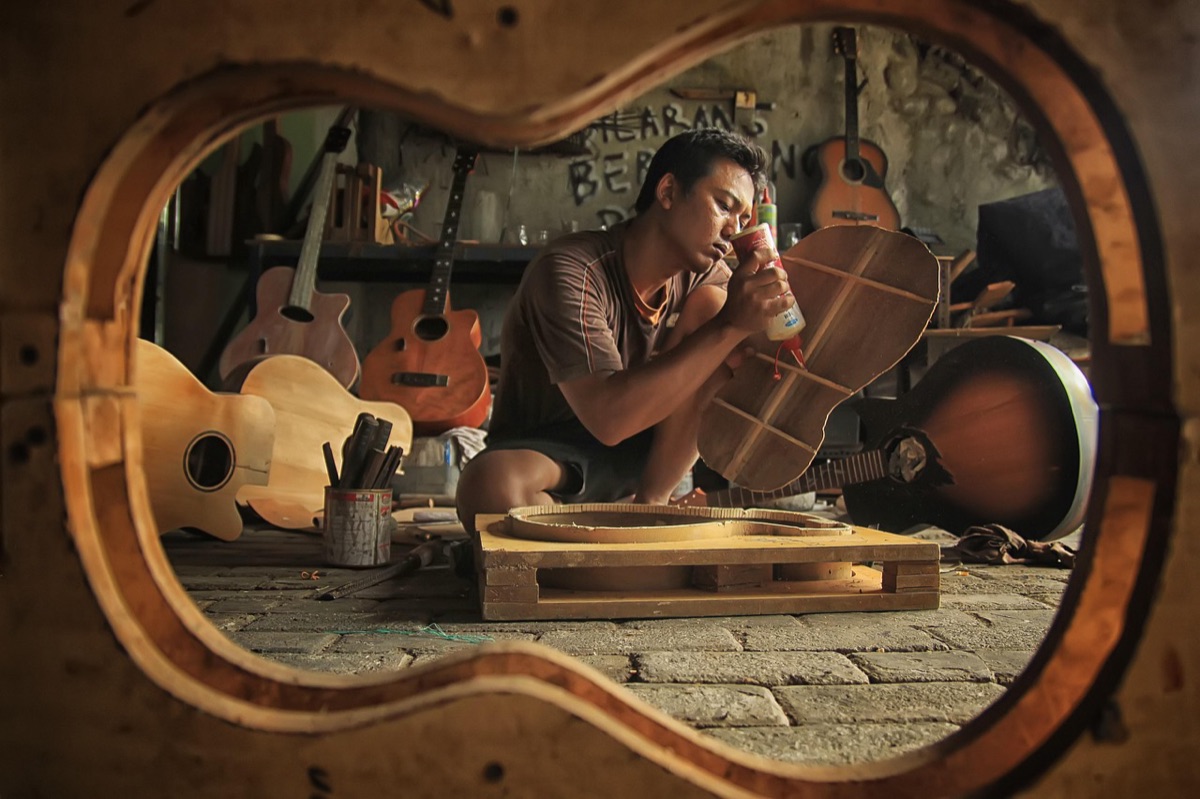 A guitar maker sanding a guitar top in his workshop, seen through a guitar body