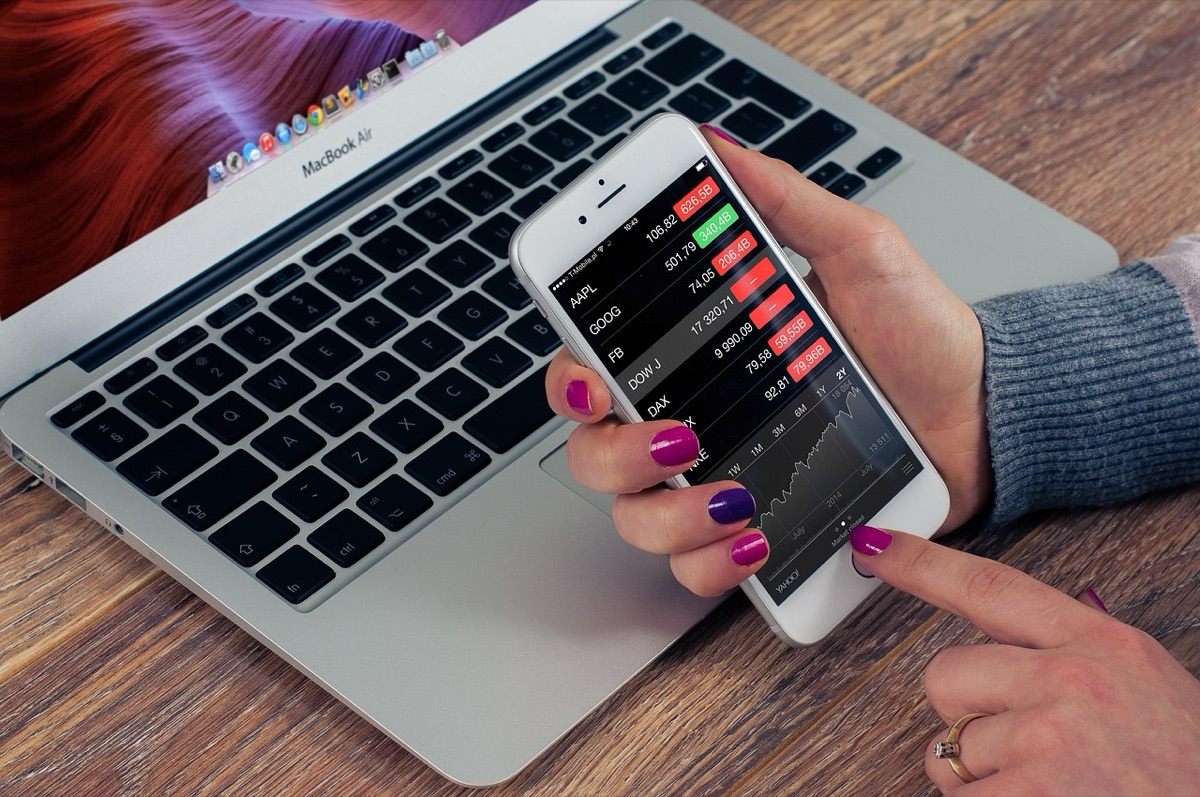 Hands holding a phone with a trading app over a laptop